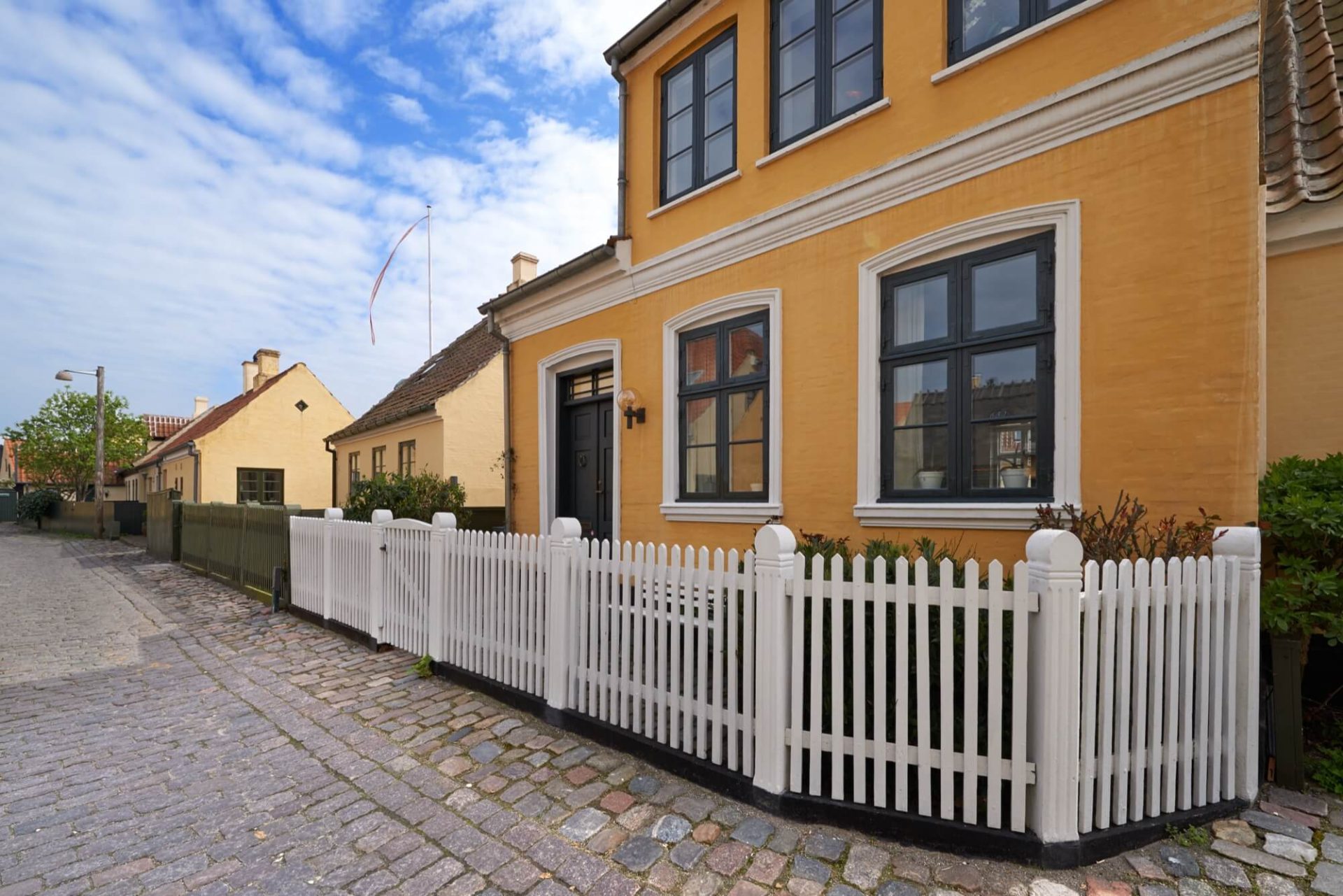 En gul putsad villa, framhävd av dess svarta fönster och omgiven av en vit trägrind, står längs en charmig kullerstensgata