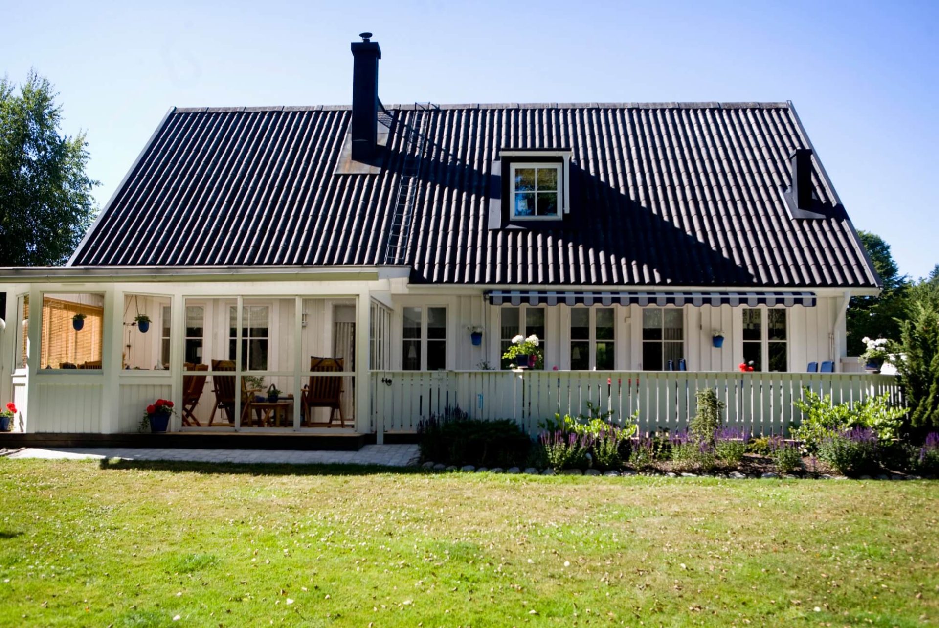 En charmfull vit trävilla, toppad med ett svart tak, omringad av en luftig veranda och en trädgård som frodas